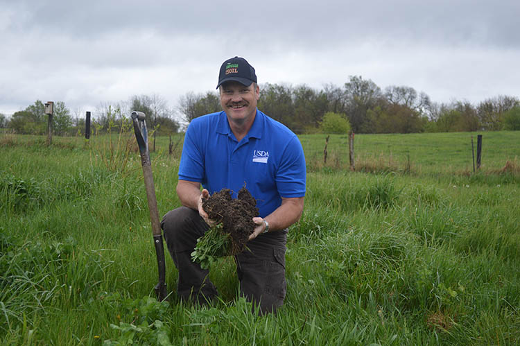 Michael Henderson, the State Agronomist for NRCS in Des Moines, IA, holding up a chunk of soil and grass in a green prairie with a shovel standing up next to him.