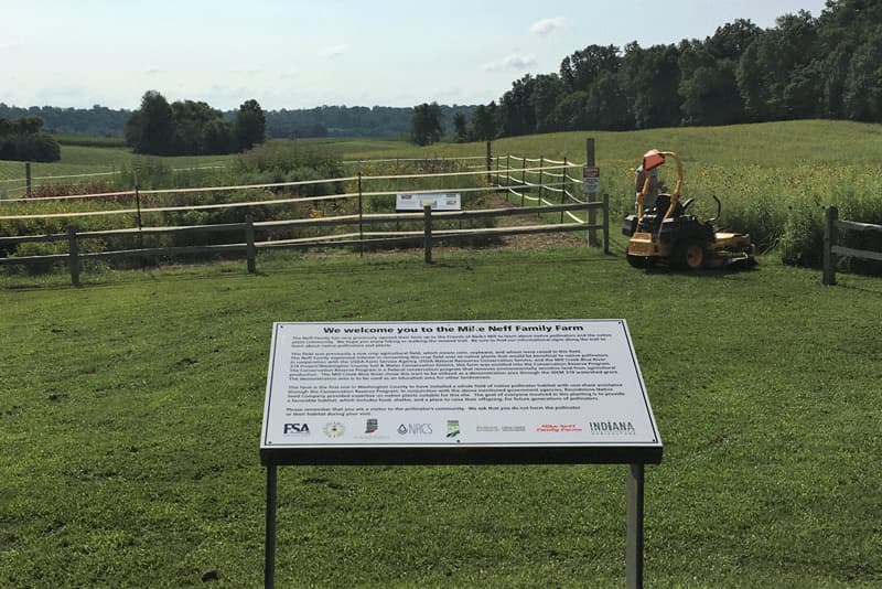 A picture of a sign in front of a fenced in area and a mower. The sign reads: "We welcome you to the Mike Neff Family Farm" followed by several illegible paragraphs and the logos of several government agencies, including FSA and NRCS