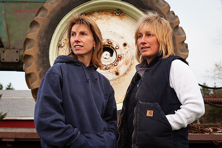 A photo of​ Nina and Kim standing in front of heavy-duty farm equipment.