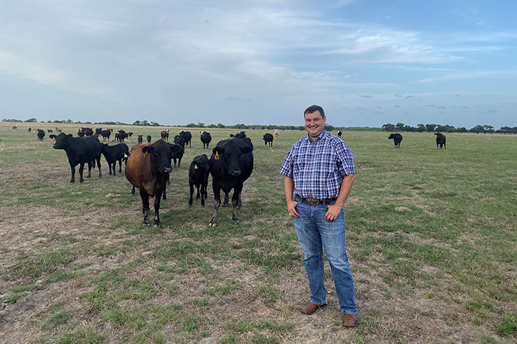 Scott Reed dressed in blue jeans and a blue plaid shirt stands in a pasture with cows in the background.