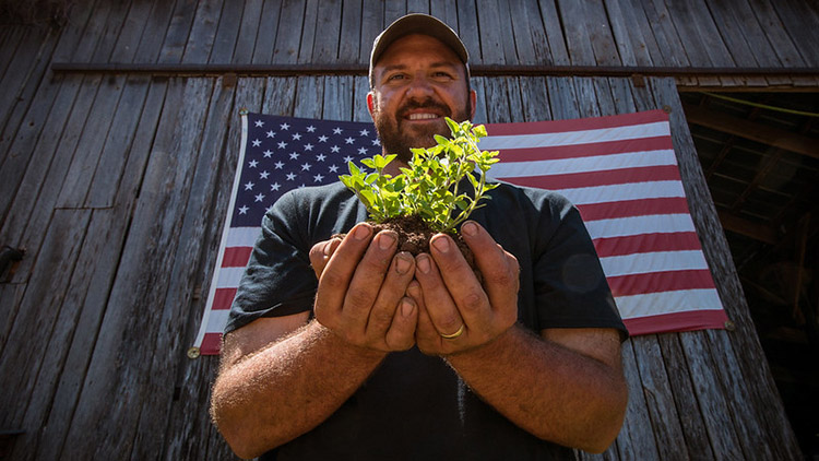 A farmer holds a plant while standing in front of an American flag. 