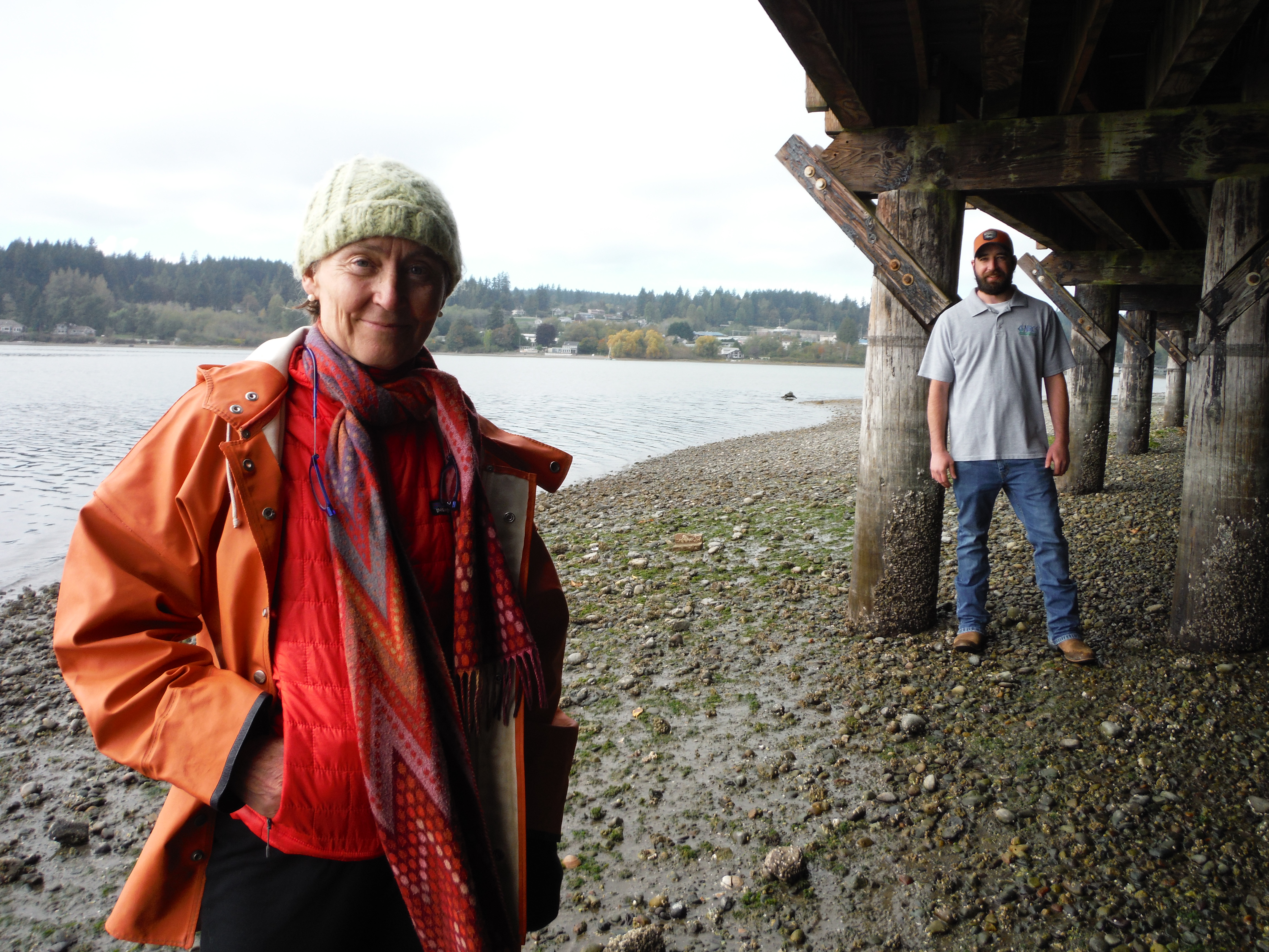 Two people stand under dock on the water's side