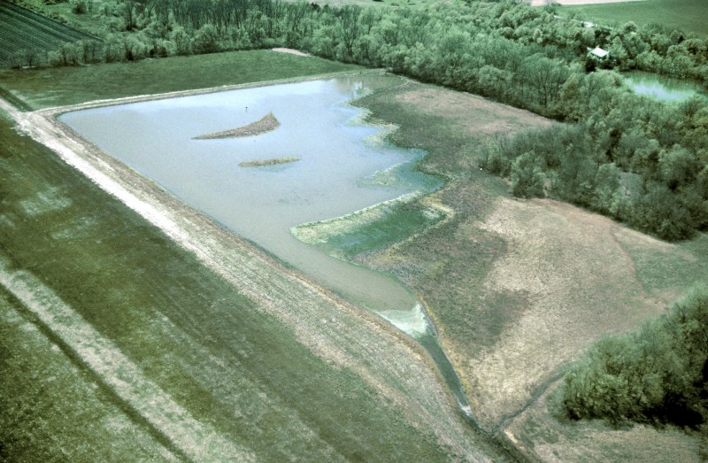 Aerial photo of a green field with water in it surrounded by trees.