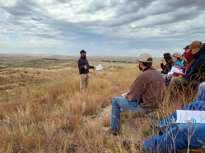 People sitting outside in brown grass listening to a speaker.