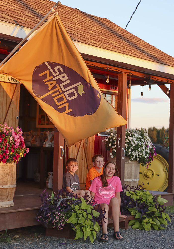 Three smiling kids sitting in front of a farmstand.