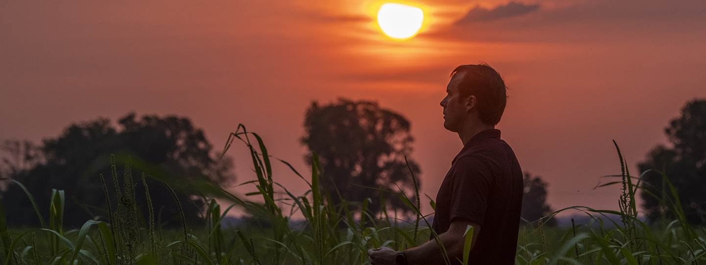 A man stands in a field against the setting sun