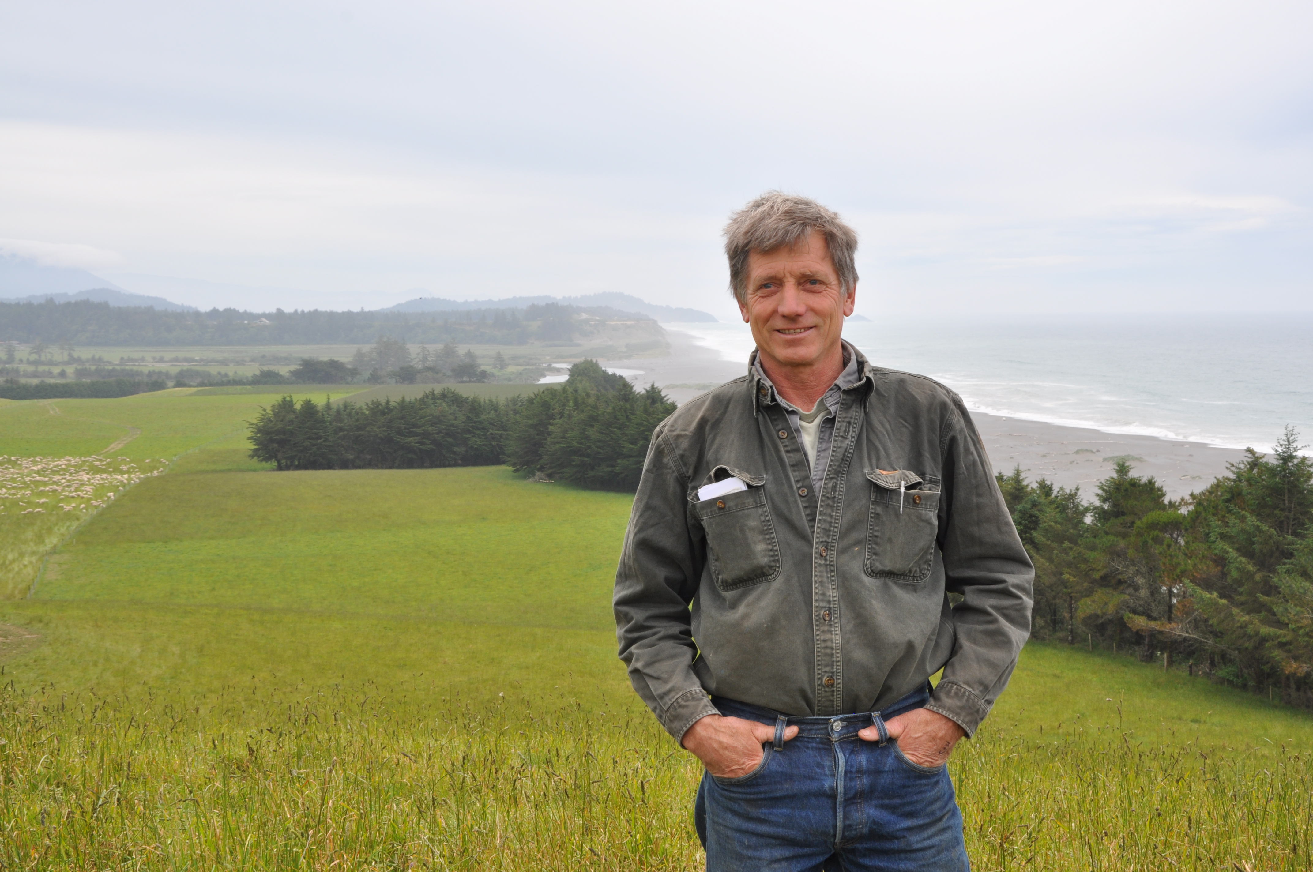 Pete Wahl poses at Wahl Family Farm as sheep graze atop cliffs overlooking the union of the Elk River and Pacific Ocean.