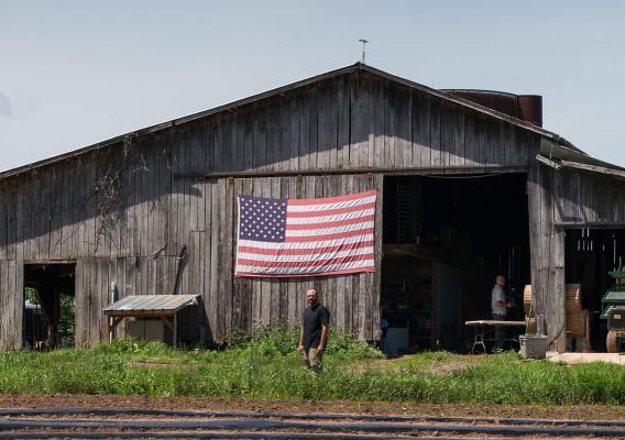 Veteran farmer standing in front of barn with an American flag behind him