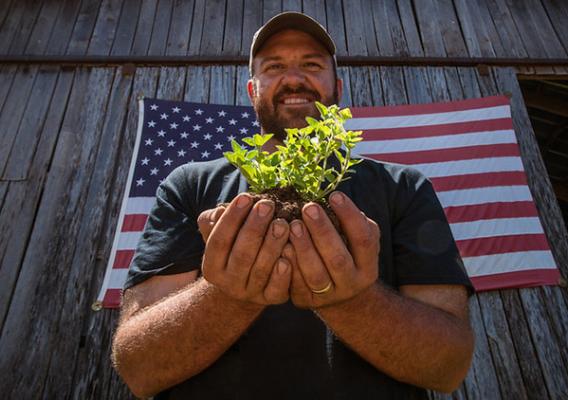 A farmer holds a plant and stands in front of an American flag. 