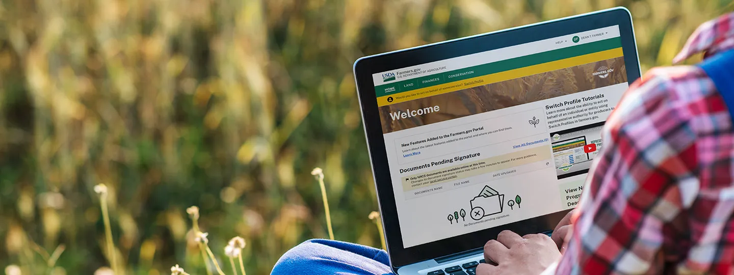 A person sits in a field with an open laptop and the browser on the farmers.gov website